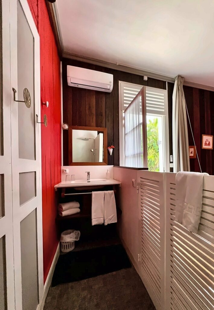 Salle d'eau de la chambre Marie : vasque, miroir et fenêtre à persiennes ouverte sur le jardin — Villa Myriam, Saint-Pierre